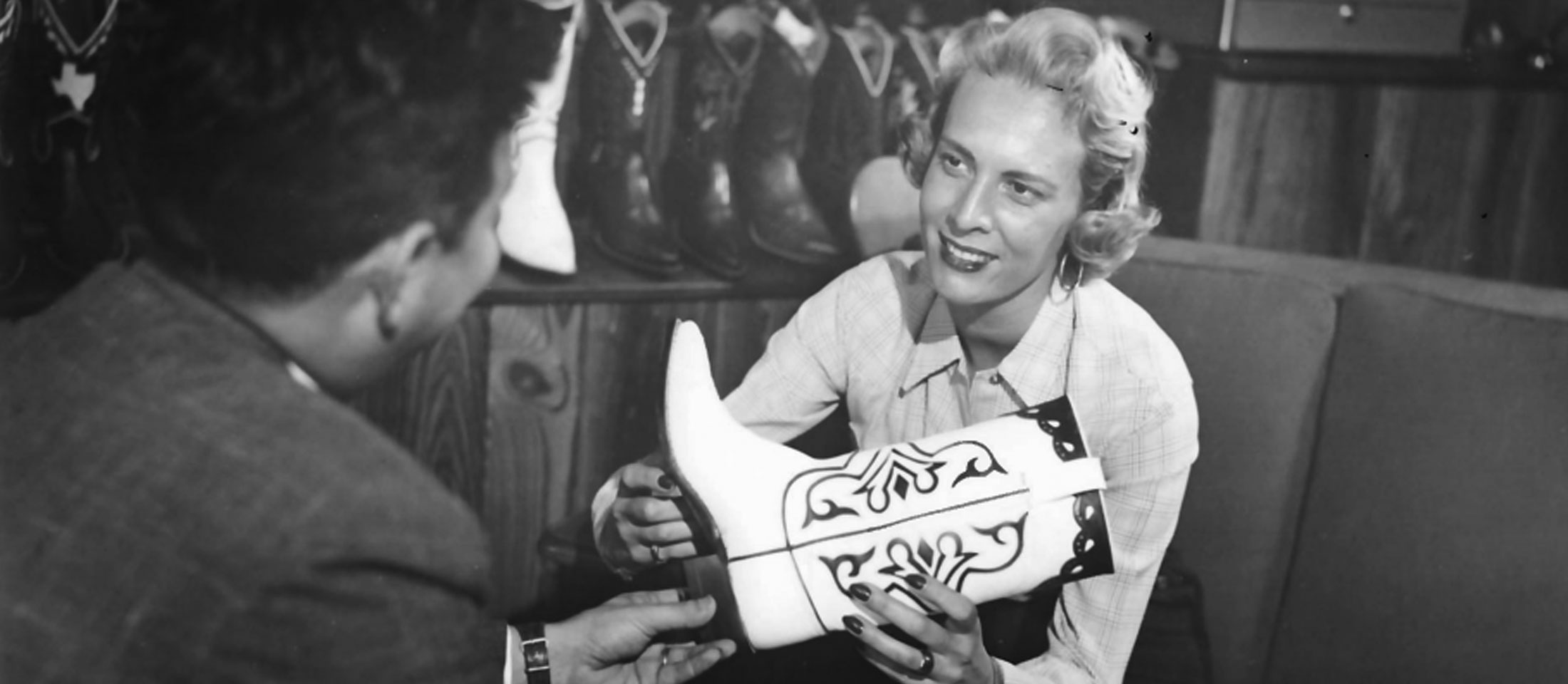 A black and white photo of a young Jane Chilton Justin with blonde hair, wearing business attire and holding a cowboy boot, presenting a boot to a customer.